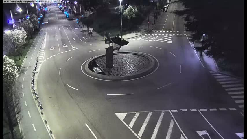 A nighttime aerial view shows a mostly empty roundabout with a dark sculpture in the center, surrounded by roads and pedestrian crossings.