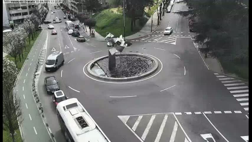 A roundabout with a modern sculpture in the center is surrounded by several cars and a bus, on a partly cloudy day.