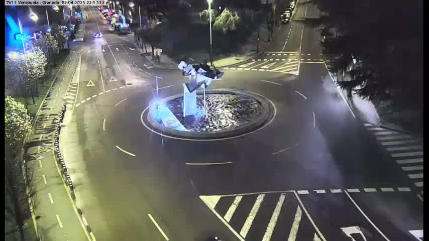 A nighttime aerial view of a roundabout with a modern sculpture in the center, surrounded by wet roads and pedestrian crossings.