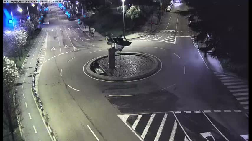 A nighttime aerial view shows a roundabout with a dark sculpture in the center, surrounded by roads with crosswalks and minimal traffic.