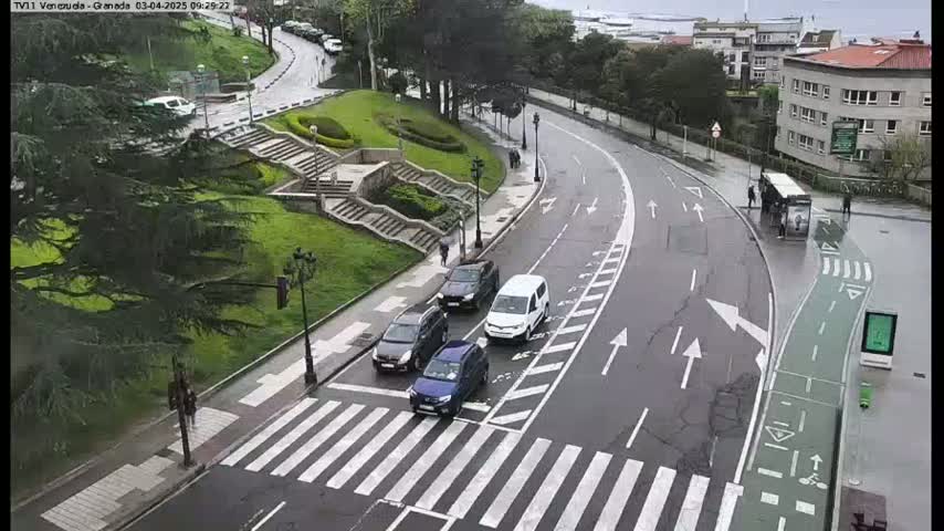 A wet, curving road with several cars and pedestrians near a pedestrian crossing and a bus stop, next to a grassy area with stairs and trees.