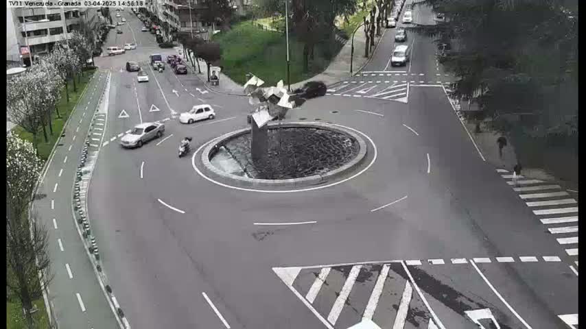 A roundabout with several cars and a person on a scooter is shown on a mostly gray asphalt road, with a modern sculpture at the center, under a cloudy sky.