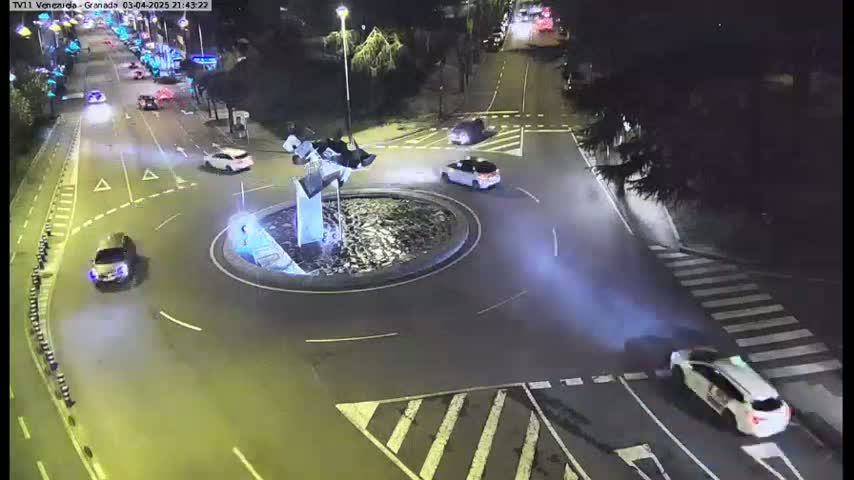 A nighttime aerial view of a roundabout with several cars driving around it and on the surrounding roads.