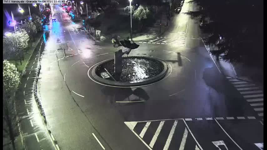 A wet roundabout at night is centered in the image, with sparsely trafficked roads extending in multiple directions.