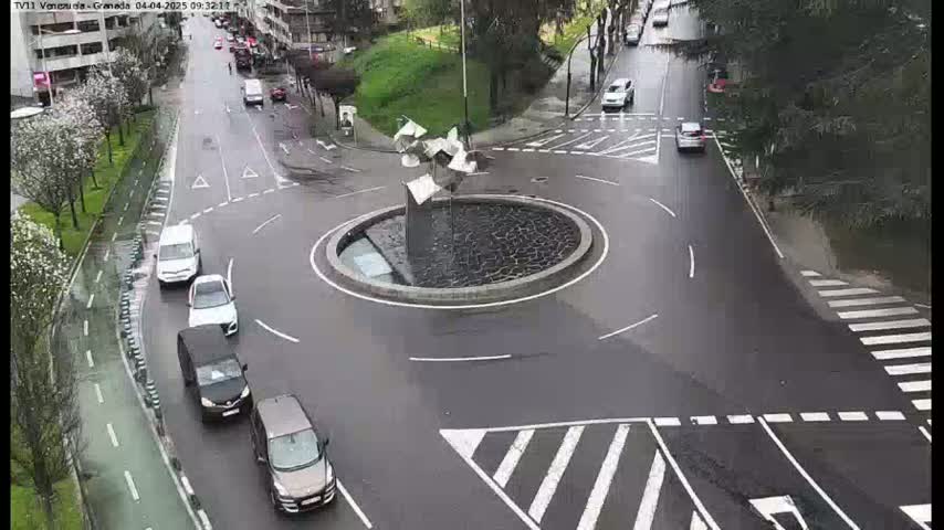 Several cars are driving around a wet roundabout featuring a modern sculpture in the center.