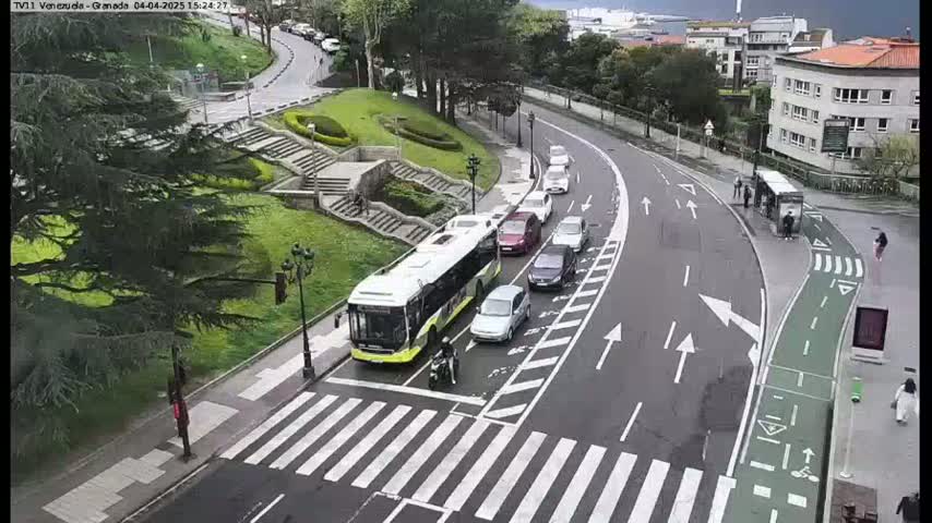 A city street scene shows a bus and several cars on a roadway that curves around a grassy area with stairs, under an overcast sky.