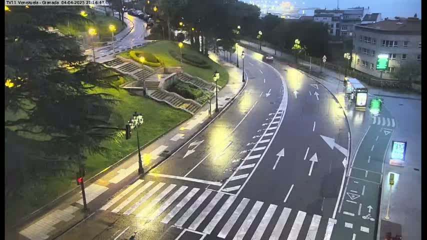 A wet, curved road with a crosswalk and bike lane is seen at night, with a few cars and a bus visible, near stairs leading to a grassy area with trees and buildings in the background.