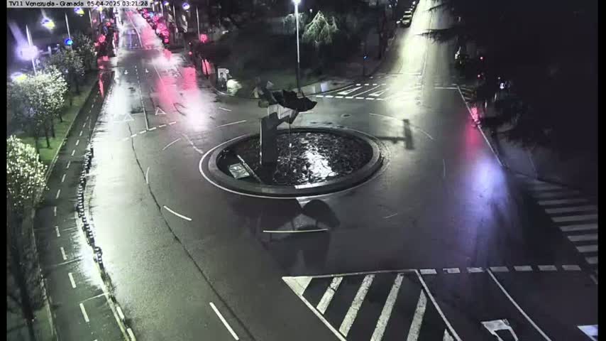 A wet roundabout with a dark sculpture at its center is surrounded by roadways with sparse nighttime traffic.