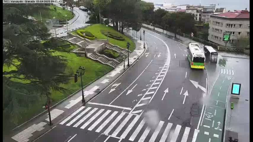 A wet road curves through a green park with stairs and two buses stopped at a bus stop, under a rainy sky.