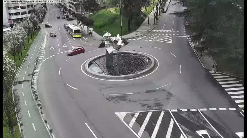 A roundabout with a modern sculpture in the center is surrounded by roads with several cars and pedestrians, on a partly cloudy day.