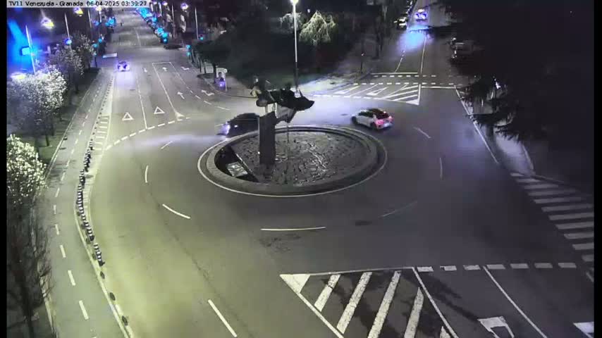 At night, several cars navigate a roundabout centered around a dark sculpture, with streetlights illuminating the asphalt roads and surrounding trees.