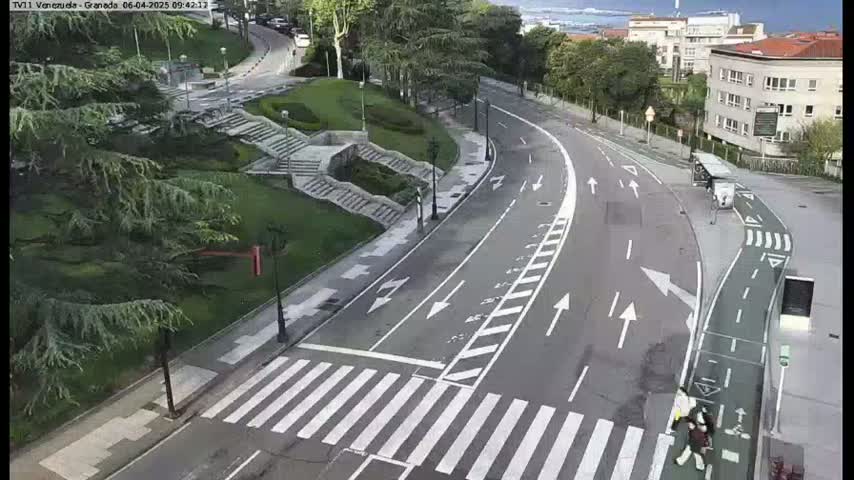 A mostly empty road curves through a city park on an overcast day, with a crosswalk and pedestrian areas visible.