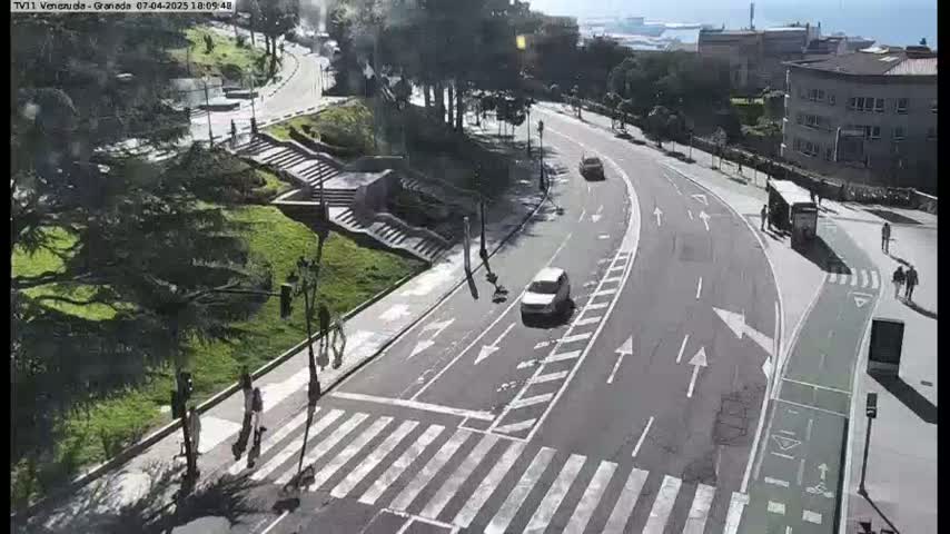 A sunny day overlooking a curving road with pedestrians and cars, alongside a grassy area with stairs and trees.