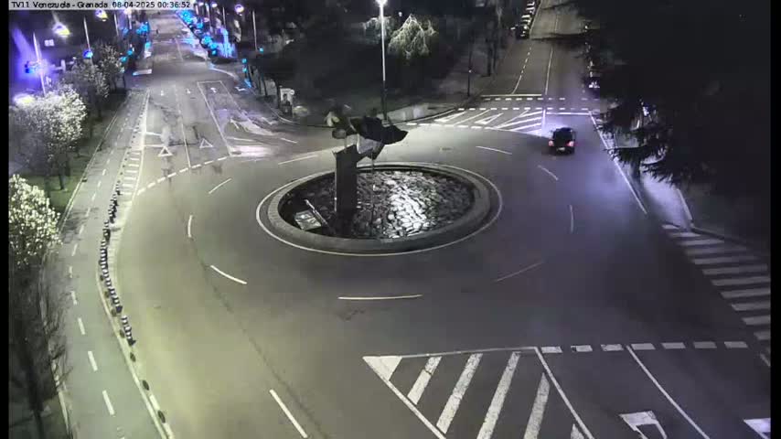 A nighttime aerial view shows a roundabout with a sculpture in the center, surrounded by roads with cars and pedestrian crosswalks.
