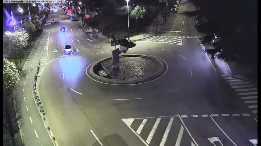 A nighttime aerial view shows a roundabout with a dark sculpture in the center, several cars driving on surrounding roads, and pedestrian crosswalks.