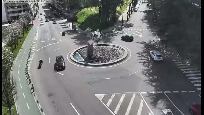 A roundabout with several cars and a motorcycle is shown on a sunny day.