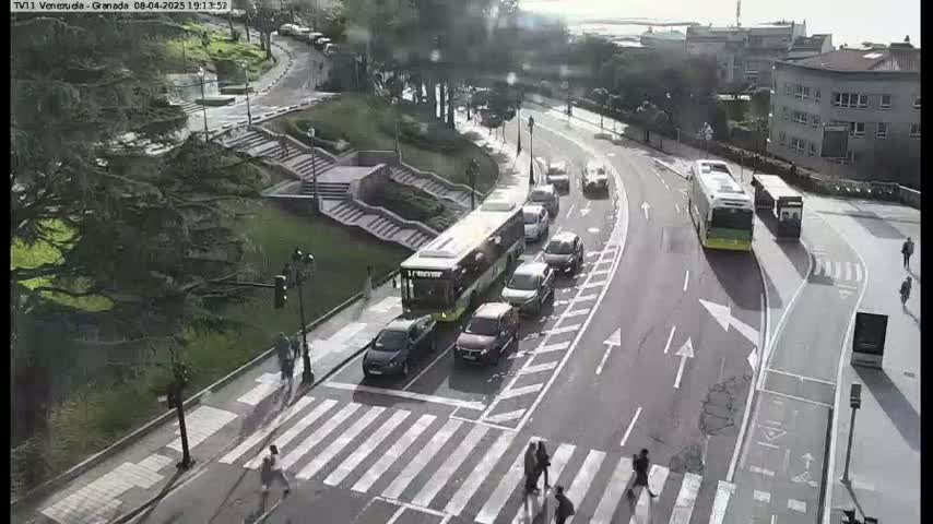 A sunny day reveals a city street scene with several cars and buses navigating a curving road near a pedestrian crossing and stairways leading up to a tree-lined hillside.