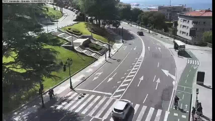 A sunny day overlooking a curved roadway with crosswalks, a bike lane, and pedestrians walking near a grassy area with stairs and some buildings in the background.
