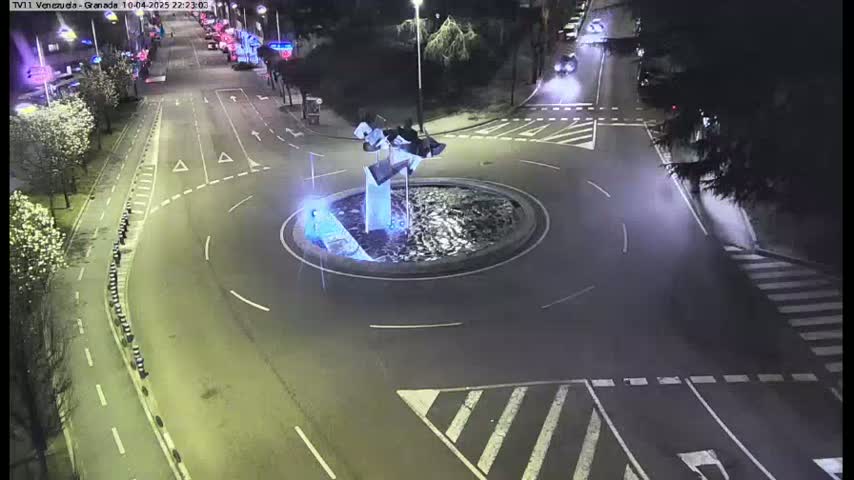 A nighttime aerial view shows a roundabout with a modern sculpture at its center, surrounded by roadways and pedestrian crossings.
