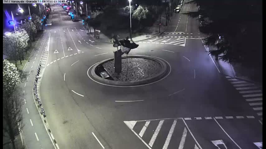 A nighttime aerial view shows a mostly empty roundabout with a dark sculpture in the center, surrounded by roads with street markings and pedestrian crosswalks.