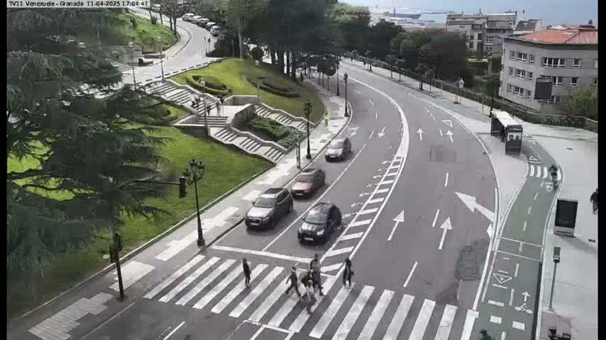 A curved road with several cars and pedestrians crossing at a crosswalk, situated in a park-like setting with stairs and lush greenery under a partly cloudy sky.