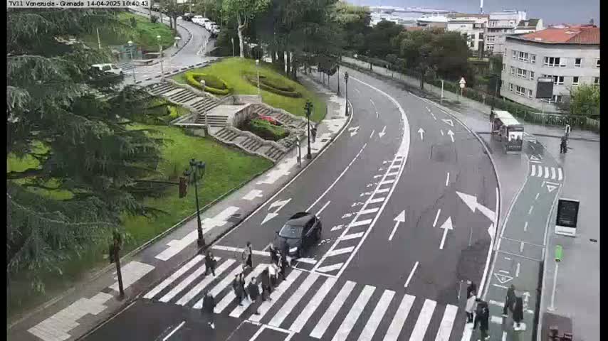 A wet street scene shows a crosswalk with pedestrians, cars, and a bus on a curved road next to a grassy area with stairs and trees.
