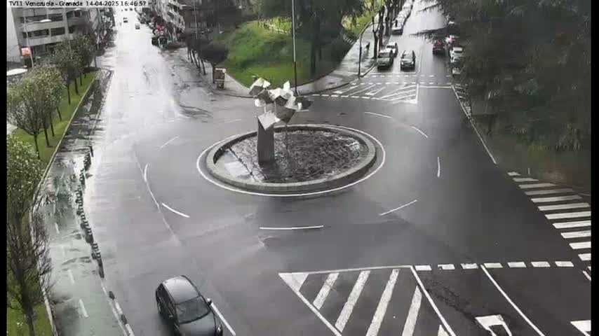 A wet roundabout with a modern sculpture in the center is surrounded by roadways with several cars and pedestrian crossings.