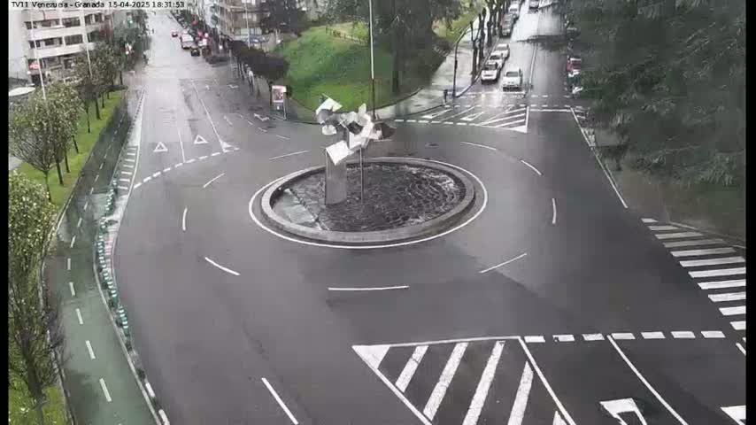 A wet roundabout with a modern sculpture in the center is surrounded by roadways and pedestrian crossings.