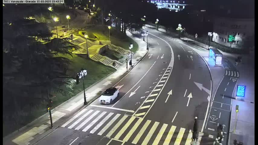 A white car is stopped at a crosswalk on a curving, well-lit road at night, with a few pedestrians visible near some stairs and buildings.