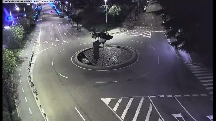 A nighttime aerial view shows a roundabout with a dark sculpture in the center, surrounded by roads with minimal traffic.