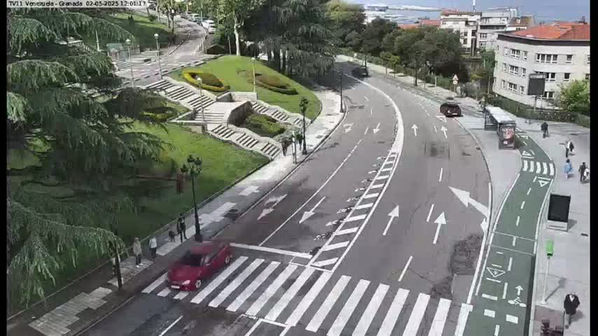 A red car crosses a pedestrian crosswalk on a curved road that winds through a green park-like area with steps and buildings in the background on a partly sunny day.