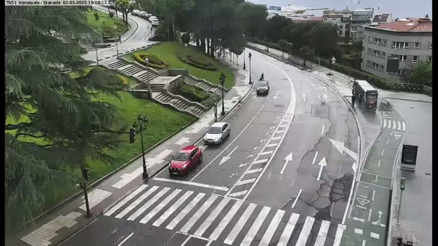 A wet, curved road with several cars and a truck is seen from an elevated viewpoint, next to a grassy area with stone steps and lush green trees.