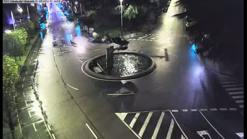 A wet, nighttime view of a roundabout with a dark sculpture in the center, surrounded by roads with sparse traffic.