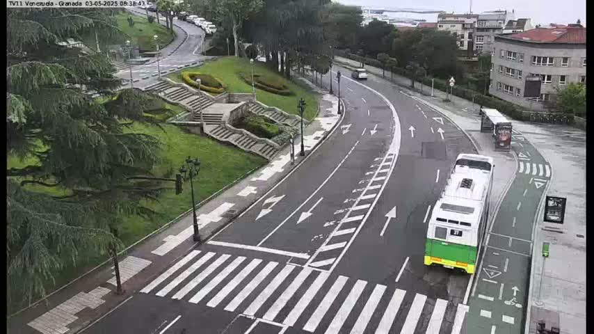 A curved road with a crosswalk and two buses, one of which is green and white, is seen on an overcast day next to a grassy area with stairs and trees.