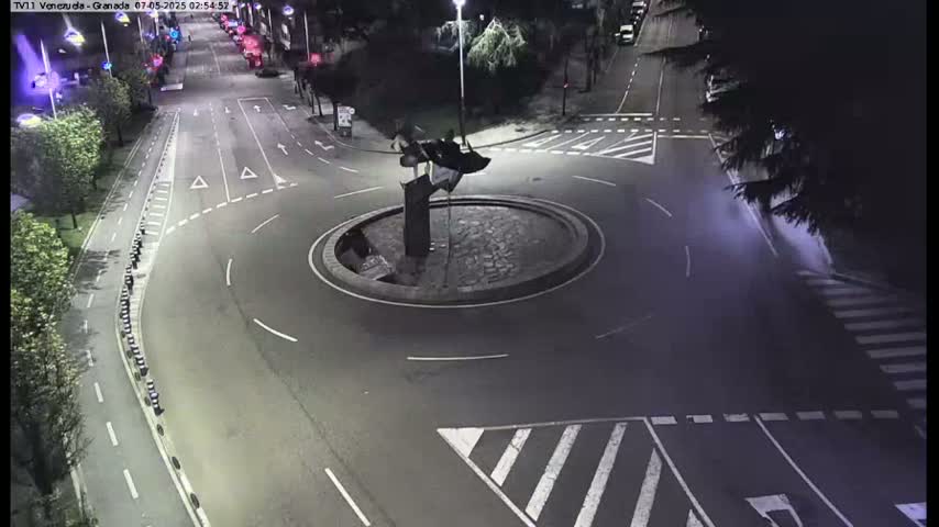 A nighttime aerial view shows a roundabout with a dark sculpture in the center, surrounded by roads with crosswalks and sparse traffic.