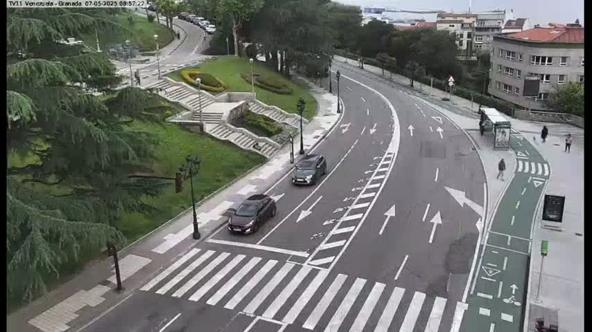 A curved road with two dark-colored cars and a pedestrian crossing, bordered by green landscaping, stairs, and buildings, on an overcast day.
