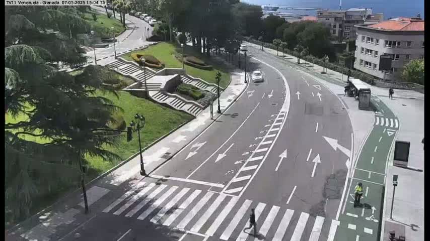 A sunny day overlooking a curved road with a crosswalk, a bike lane, and a landscaped area with stairs, trees, and a few pedestrians.