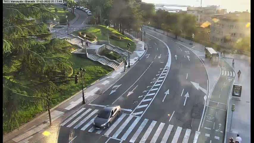 A sunny, high-angle view shows a curving road with a crosswalk and bicycle lane, alongside a park with stairs and lush green trees.