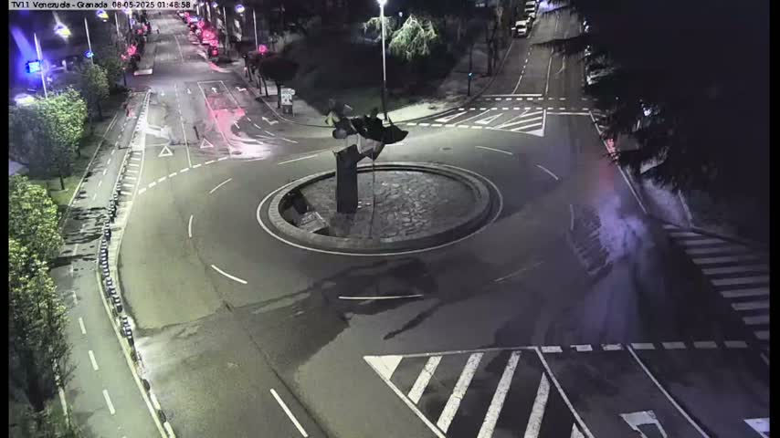 A nighttime aerial view of a roundabout with a sculpture in the center, surrounded by wet roads and pedestrian crossings.