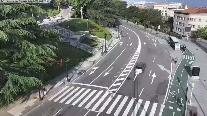 A sunny day overlooking a roadway with a crosswalk and bike lane, next to a park with stairs and lush green trees.