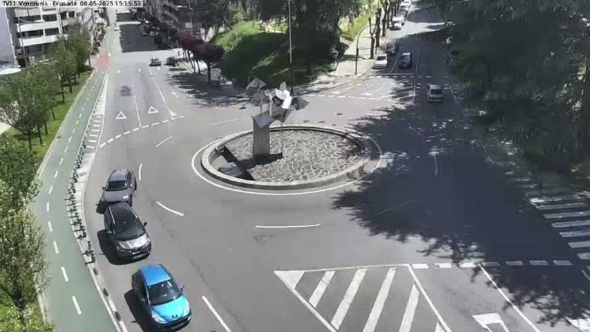 A sunny overhead view shows a traffic circle with several cars, a bike lane, and a modern, metallic sculpture in the center.
