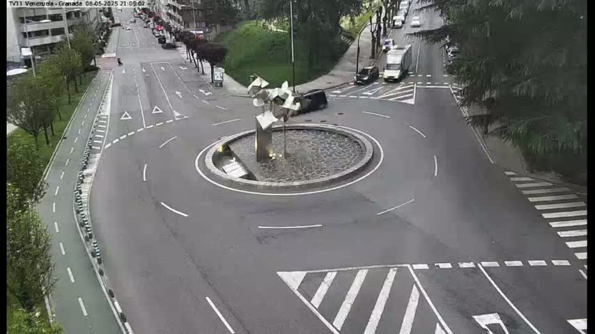 A traffic roundabout with a modern sculpture in the center is situated on a road with bike lanes and pedestrian crosswalks, under overcast conditions.