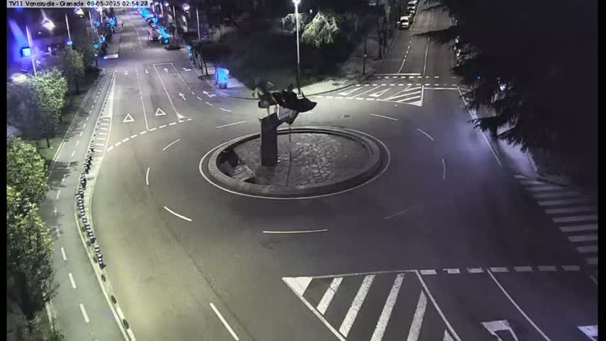 A nighttime aerial view shows a roundabout with a dark sculpture in the center, surrounded by roads with minimal traffic.