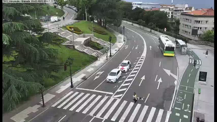 A curved road with two white cars and a bus, a pedestrian crossing, and a bike lane next to a grassy area with stairs and trees, under an overcast sky.