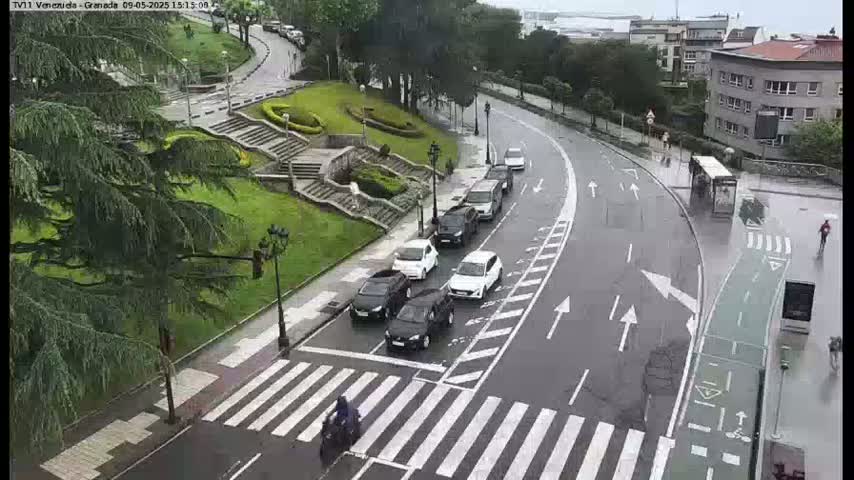 A wet, city street scene shows several cars stopped at a crosswalk near a staircase leading up to a park with lush green grass and trees.
