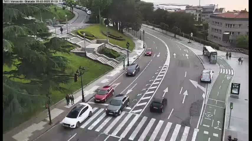 A street scene shows several cars driving on a curving road that passes by a pedestrian crossing near a grassy area with stairs and trees, under a mostly clear sky.