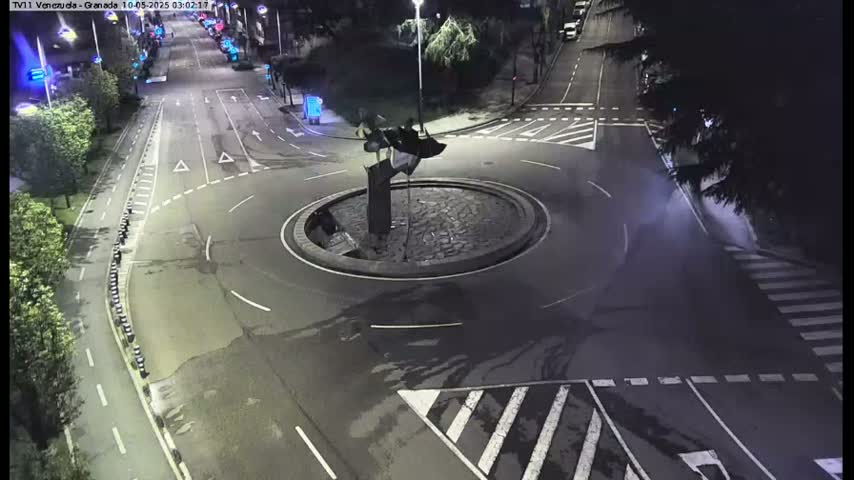 A nighttime aerial view shows a roundabout with a dark sculpture in the center, surrounded by empty roads and pedestrian crossings.
