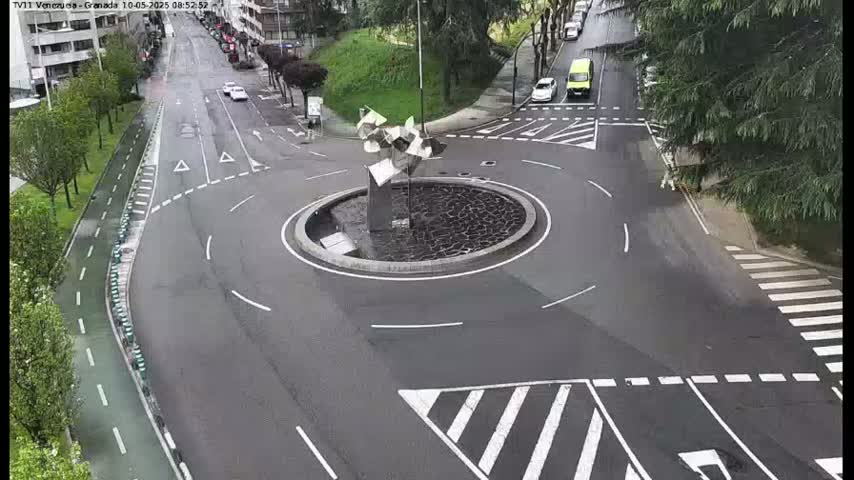 A wet roundabout with a modern sculpture in the center is surrounded by roads and a pedestrian crossing, with several cars visible.