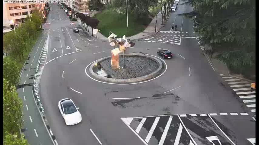 A roundabout with a modern sculpture in the center, surrounded by roads with several cars, under a partly cloudy sky.