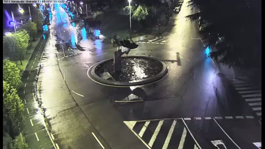 A wet roundabout with a sculpture at its center is situated at a nighttime intersection, with vehicles visible on the roads.
